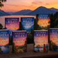 **alt-tekst:** Stapeltje boeken uit de Zeven Zussen-serie van Lucinda Riley met een kopje thee ernaast op een houten tafel